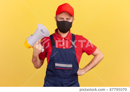 Male worker in uniform and with protective mask on face holding megaphone looking at camera with serious expression, warning about quarantine. Indoor studio shot isolated on yellow background Male worker in uniform and with protective mask on face holding megaphone looking at camera with serious expression, warning about quarantine. Indoor studio shot isolated on yellow background 87795070