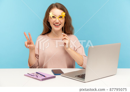 Funny optimistic woman employee covering eyes with bright paper glasses, showing victory gesture and wearing humorous masquerade accessory, having fun at workplace while working in office. indoor Funny optimistic woman employee covering eyes with bright paper glasses, showing victory gesture and wearing humorous masquerade accessory, having fun at workplace while working in office. indoor 87795075
