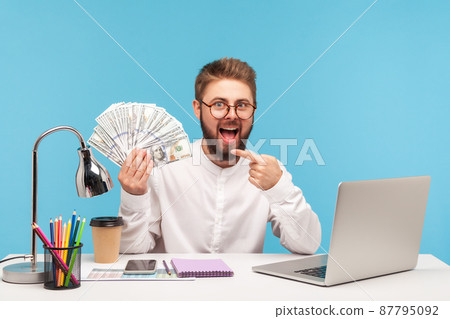 Excited bearded man office worker pointing finger at dollar cash in his hand looking at camera with big eyes and open mouth, money laundering. Indoor studio shot isolated on blue background Excited bearded man office worker pointing finger at dollar cash in his hand looking at camera with big eyes and open mouth, money laundering. Indoor studio shot isolated on blue background 87795092