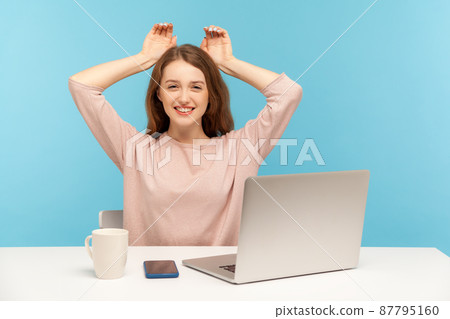 Charming young woman employee smiling and showing bunny ears on head, resting, having fun at workplace in home office, feeling carefree and happy. indoor studio shot isolated on blue background Charming young woman employee smiling and showing bunny ears on head, resting, having fun at workplace in home office, feeling carefree and happy. indoor studio shot isolated on blue background 87795160