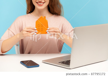 Closeup of young positive woman, real estate agent sitting at workplace with laptop and holding small paper house, smiling to camera, offering rental and home purchase. indoor studio shot isolated 87795172