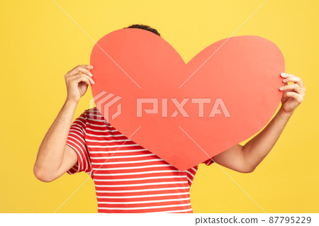 Unknown man in red striped t-shirt hiding his face behind big red paper heart, making anonymous surprise, sending greeting card. Indoor studio shot isolated on yellow background 87795229
