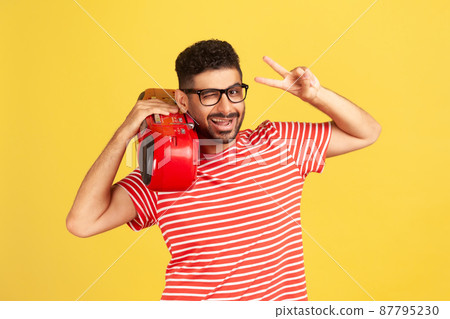 Smiling cheerful man with beard in eyeglasses and striped t-shirt holding record player on shoulder and showing victory gesture with fingers. Indoor studio shot isolated on yellow background Smiling cheerful man with beard in eyeglasses and striped t-shirt holding record player on shoulder and showing victory gesture with fingers. Indoor studio shot isolated on yellow background 87795230
