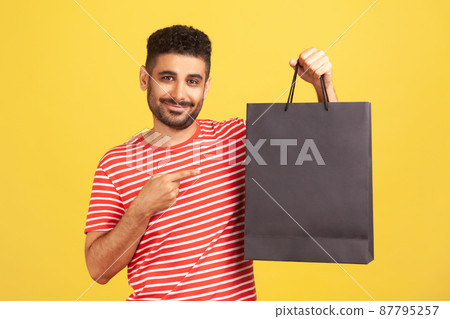 Smiling bearded man in striped t-shirt pointing finger at black paper bag with purchases, satisfied with shopping, seasonal sale. Indoor studio shot isolated on yellow background Smiling bearded man in striped t-shirt pointing finger at black paper bag with purchases, satisfied with shopping, seasonal sale. Indoor studio shot isolated on yellow background 87795257