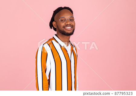 Happy bearded afro-american man with dreadlocks in stylish striped shirt and eyeglasses looking at camera and smiling, optimism. Indoor studio shot isolated on pink background Happy bearded afro-american man with dreadlocks in stylish striped shirt and eyeglasses looking at camera and smiling, optimism. Indoor studio shot isolated on pink background 87795323