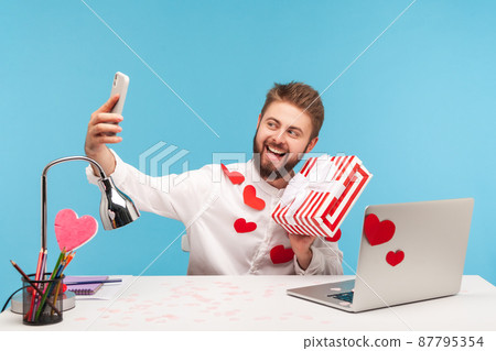 Cheerful positive man blogger with beard in white shirt covered with heart shaped stickers holding and showing on smartphone camera striped giftbox. Indoor studio shot isolated on blue background Cheerful positive man blogger with beard in white shirt covered with heart shaped stickers holding and showing on smartphone camera striped giftbox. Indoor studio shot isolated on blue background 87795354