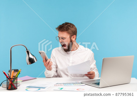 Angry nervous man office worker in white shirt screaming looking at smartphone display, skipping important event through lot of paper work. Indoor studio shot isolated on blue background Angry nervous man office worker in white shirt screaming looking at smartphone display, skipping important event through lot of paper work. Indoor studio shot isolated on blue background 87795361