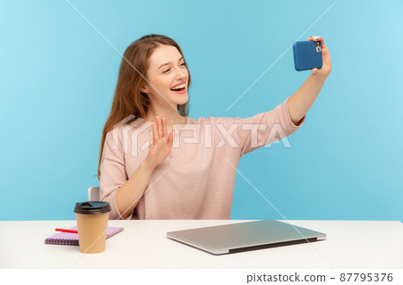 Optimistic friendly woman, office employee or blogger, sitting at workplace and taking selfie by mobile phone, talking on video call and saying hello hi, waving hand. indoor studio shot isolated Optimistic friendly woman, office employee or blogger, sitting at workplace and taking selfie by mobile phone, talking on video call and saying hello hi, waving hand. indoor studio shot isolated 87795376
