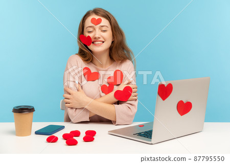 Charming happy young woman with kind smile sitting at workplace office, all covered with sticker love hearts, embracing herself, enjoying valentine's day greetings. indoor studio shot, isolated 87795550