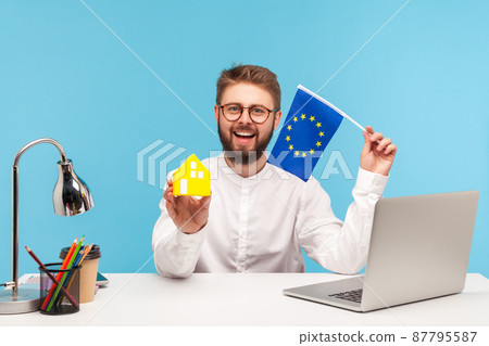 Smiling positive man real estate agency worker in eyeglasses holding paper toy house and flag of europe union, registration of visas and passports. Indoor studio shot isolated on blue background 87795587