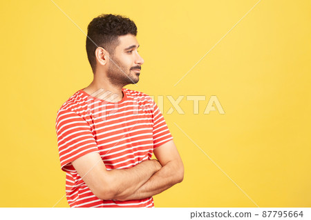 Profile portrait healthy self confident man with beard in red striped t-shirt standing folding hands on chest and looking far away. Indoor studio shot isolated on yellow background 87795664