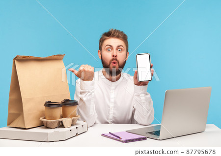 Surprised excited man pointing at fast food order on his workplace desk with thumbs and holding smartphone with empty display, using app making order. Indoor studio shot isolated on blue background 87795678