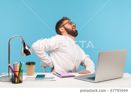 Man with beard in white shirt and eyeglasses massaging lower back sitting at workplace with laptop, feeling pain, sedentary lifestyle, stones in kidneys. Indoor studio shot isolated on blue background Man with beard in white shirt and eyeglasses massaging lower back sitting at workplace with laptop, feeling pain, sedentary lifestyle, stones in kidneys. Indoor studio shot isolated on blue background 87795694