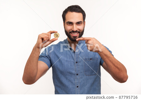 Positive cheerful man pointing finger at golden coin in his hand, advertising of btc crypto coin, looking at camera with toothy smile. Indoor studio shot isolated on white background 87795716