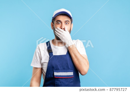 I won't say anyone. Scared handyman in overalls and gloves covering mouth with hand, afraid to talk, looking intimidated and frightened. Profession of service industry, courier delivery. isolated 87795727