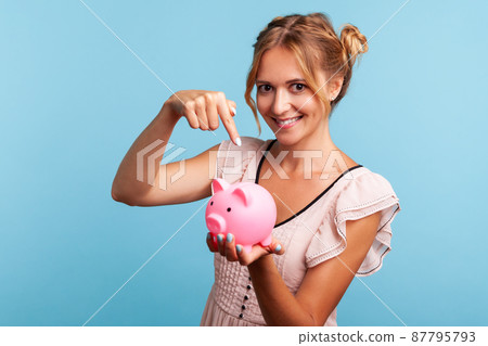 Beautiful young adult woman in summer dress holding piggy bank and pointing with index finger, suggesting you to put coins inside, saving money. Indoor studio shot isolated on blue background. 87795793