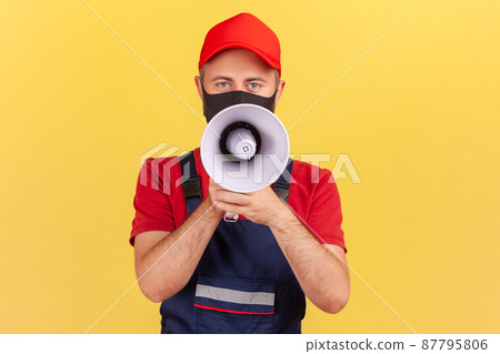 Craftsman in uniform and facial protective mask speaking holding loudspeaker near mouth, talking about safety rules on quarantine. Indoor studio shot isolated on yellow background Craftsman in uniform and facial protective mask speaking holding loudspeaker near mouth, talking about safety rules on quarantine. Indoor studio shot isolated on yellow background 87795806