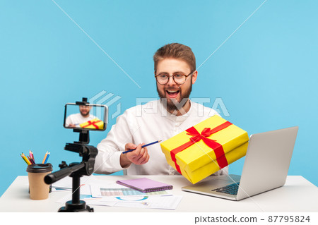Smiling man blogger holding yellow gift box with red ribbon, recording video congratulation on thankfulness for subscribers, presents packing tutorial. Indoor studio shot isolated on blue background 87795824