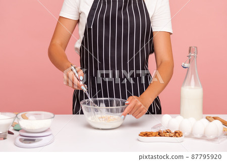 Cropped image of baker in black striped apron mixing dough with corolla, making bakery, homemade pastry, confectioner preparing cake. Indoor studio shot isolated on pink background. 87795920