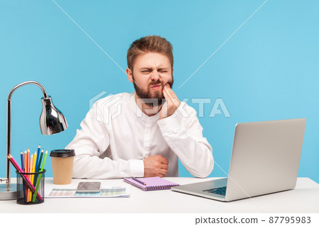 Unhappy bearded man office worker touching cheek with fingers feeling unpleasant toothache, suffering gums inflammation, dentistry problems. Indoor studio shot isolated on blue background 87795983