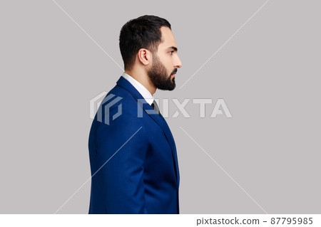 Side view portrait of confident bearded male looking at camera with serious expression, unsmiling determined business man, wearing official style suit. Indoor studio shot isolated on gray background. Side view portrait of confident bearded male looking at camera with serious expression, unsmiling determined business man, wearing official style suit. Indoor studio shot isolated on gray background. 87795985