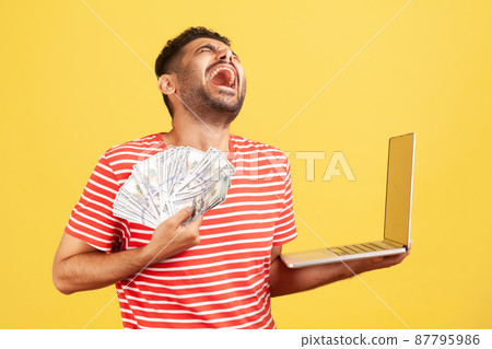 Extremely happy excited man in striped t-shirt screaming and laughing holding laptop and lot of dollar cash, enjoying jackpot winning. Indoor studio shot isolated on yellow background Extremely happy excited man in striped t-shirt screaming and laughing holding laptop and lot of dollar cash, enjoying jackpot winning. Indoor studio shot isolated on yellow background 87795986