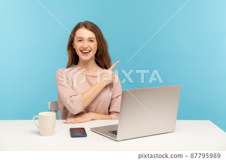 Look at advertisement. Cheerful young woman sitting at office workplace with laptop and pointing aside, showing copy space for promotional text, commercial product. indoor studio shot, isolated 87795989