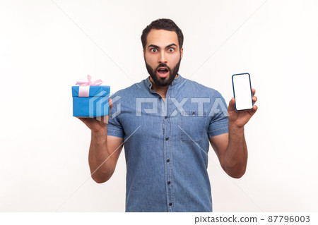 Excited surprised man with beard holding gift box and smartphone with empty screen looking at camera with big eyes, shocked with present. Indoor studio shot isolated on white background 87796003