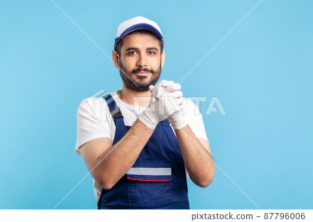 Optimistic worker in overalls and gloves keeping hands together in connection and agreement gesture. Profession of service industry, courier delivery, housekeeping maintenance. studio shot isolated 87796006
