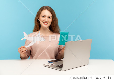 Friendly woman, travel agent holding passport and paper airplane, looking at camera with toothy smile, advertising hot tours with airline transportation. indoor studio shot isolated on blue background 87796014