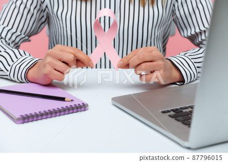 Close up female hands holding pink ribbon, symbol of breast cancer awareness, insurance company worker working on laptop making notes. Indoor studio shot isolated on pink background 87796015