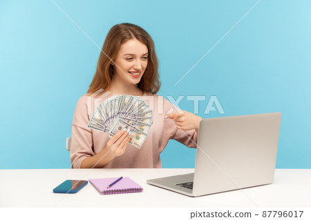 Look at money profit. Rich happy woman pointing at dollar banknotes while talking on video call on laptop, advertising online earnings, high salary. indoor studio shot isolated on blue background Look at money profit. Rich happy woman pointing at dollar banknotes while talking on video call on laptop, advertising online earnings, high salary. indoor studio shot isolated on blue background 87796017