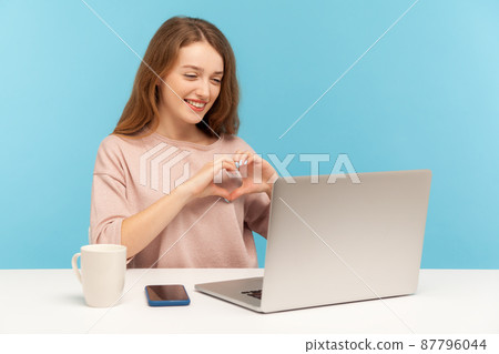 Online dating. Beautiful kind woman making heart shape with hands, expressing love and looking at laptop screen, talking on video call from home office. indoor studio shot isolated on blue background Online dating. Beautiful kind woman making heart shape with hands, expressing love and looking at laptop screen, talking on video call from home office. indoor studio shot isolated on blue background 87796044