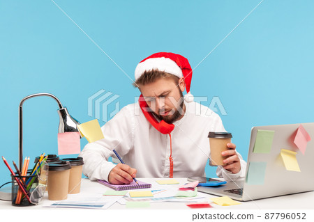 Man in santa claus hat sitting at workplace drinking coffee and talking landline telephone, writing down information into notepad, working on holidays. Indoor studio shot isolated on blue background Man in santa claus hat sitting at workplace drinking coffee and talking landline telephone, writing down information into notepad, working on holidays. Indoor studio shot isolated on blue background 87796052