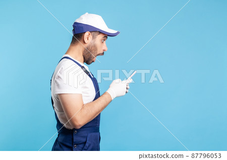Side view, surprised workman in overalls and gloves reading message on mobile phone, shocked by online delivery order app, house repair maintenance services. indoor studio shot isolated on blue Side view, surprised workman in overalls and gloves reading message on mobile phone, shocked by online delivery order app, house repair maintenance services. indoor studio shot isolated on blue 87796053
