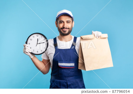 Food delivery, shipping on time. Portrait of handyman in uniform, gloves holding clock and parcel. Express courier transportation, post mail services. indoor studio shot isolated on blue background Food delivery, shipping on time. Portrait of handyman in uniform, gloves holding clock and parcel. Express courier transportation, post mail services. indoor studio shot isolated on blue background 87796054