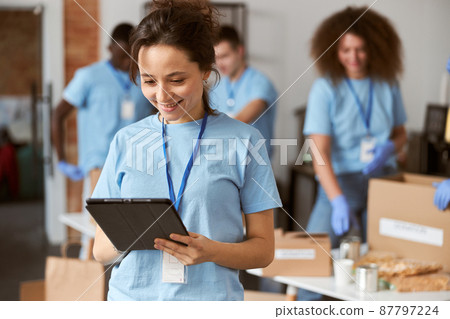 Portrait of charming young female volunteer in blue uniform using tablet pc and smiling while standing indoors. Team sorting, packing items in the background 87797224
