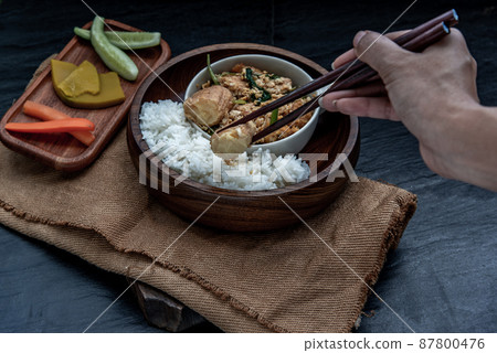 The hand is using chopsticks to pick up Stir fried egg tofu with spring onion with Herbs vegetables served with steamed rice in wooden bowl. The hand is using chopsticks to pick up Stir fried egg tofu with spring onion with Herbs vegetables served with steamed rice in wooden bowl. 87800476