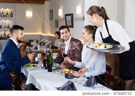 Polite smiling waitress bringing ordered dishes to guests at restaurant 87800732