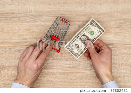 Businessman pushes the toy cart and holds one dollar bill in the hand Businessman pushes the toy cart and holds one dollar bill in the hand 87802481