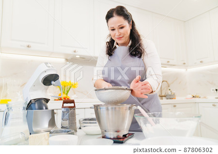 A woman cook sifts the flour through a sieve to prepare dough in a mixer bowl. 87806302