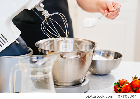 A cook adds a spoonful of flour to prepare dough in a mixer bowl. A cook adds a spoonful of flour to prepare dough in a mixer bowl. 87806304