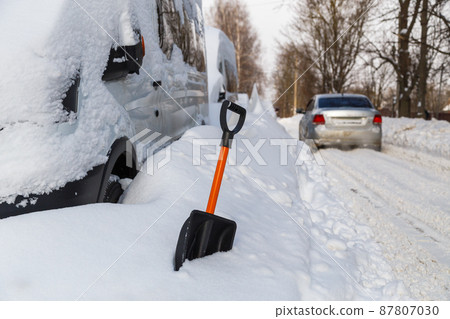 Plastic snow shovel in front of snow-covered minivan at sunny winter morning and silver car passing by in blurry background 87807030