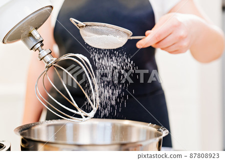 A cook sifts the powdered sugar through a sieve to prepare dough in a mixer bowl A cook sifts the powdered sugar through a sieve to prepare dough in a mixer bowl 87808923