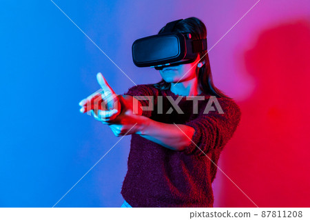 Woman with virtual reality glasses on head playing shooter game, pointing finger gun and shooting right on target, killing in cyberspace using VR headset technology. colorful neon light studio shot Woman with virtual reality glasses on head playing shooter game, pointing finger gun and shooting right on target, killing in cyberspace using VR headset technology. colorful neon light studio shot 87811208