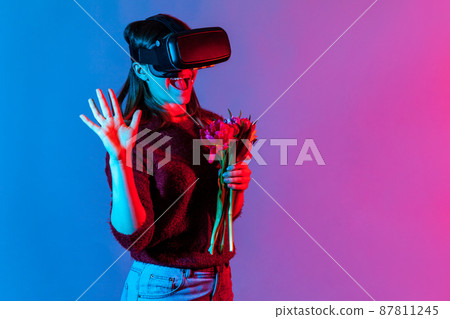 Joyful girl in VR glasses headset holding flowers bouquet and greeting virtual friend with hello hi gesture, playing date in cyberspace augmented reality, romantic relations. neon light studio shot Joyful girl in VR glasses headset holding flowers bouquet and greeting virtual friend with hello hi gesture, playing date in cyberspace augmented reality, romantic relations. neon light studio shot 87811245