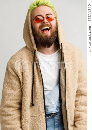 Handsome young adult bearded model man wearing beige jacket looking at camera and laughing out loud, expressing positive and optimism. Indoor studio shot isolated over gray background. 87811249