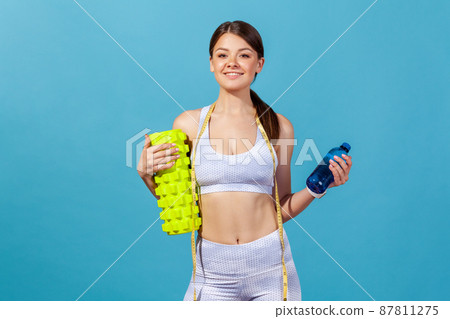 Slim self confident woman in white sportswear holding fitness roller and bottle with fresh table water, going on training at gym. Indoor studio shot isolated on blue background 87811275