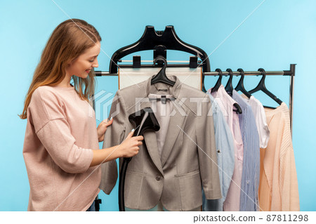 Cheerful housewife steaming business jacket at dry-cleaner and looking at fabric, advertisement of professional services washing, cleaning and ironing garment. indoor studio shot, blue background Cheerful housewife steaming business jacket at dry-cleaner and looking at fabric, advertisement of professional services washing, cleaning and ironing garment. indoor studio shot, blue background 87811298