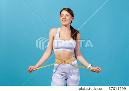 Slim athletic woman in white sports tights and top crossing tape line on her waist, measuring centimeters of body, keeping healthy lifestyle, workout. Indoor studio shot isolated on blue background Slim athletic woman in white sports tights and top crossing tape line on her waist, measuring centimeters of body, keeping healthy lifestyle, workout. Indoor studio shot isolated on blue background 87811299